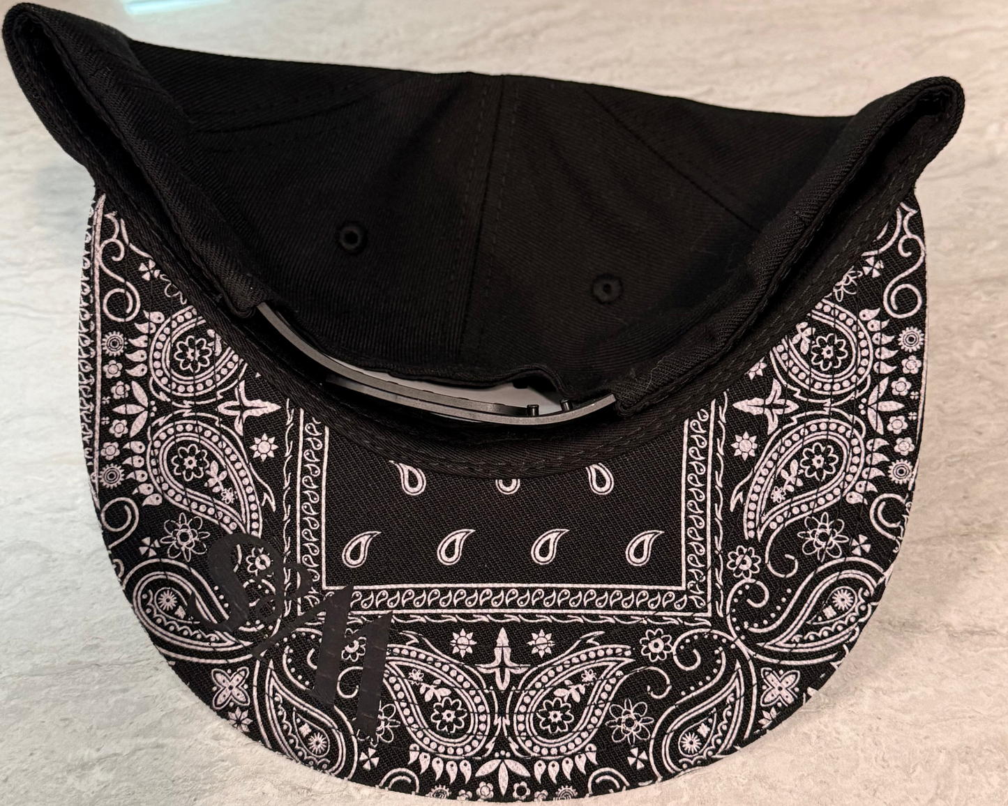 Underneath a black snapback with a black and white paisley bill and a logo W&S in black on the bill on a neutral table background