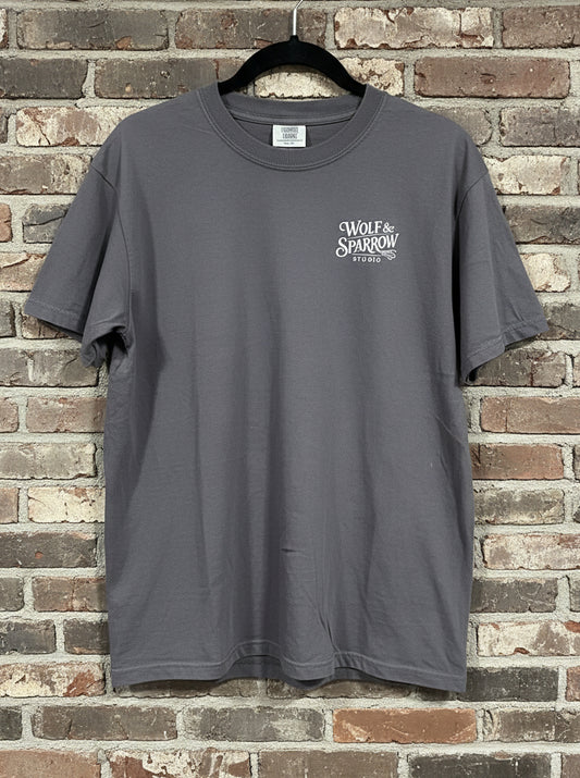 Front of a graphite t-shirt with a white logo on the breast that says "Wolf & Sparrow Studio" against a grey brick background
