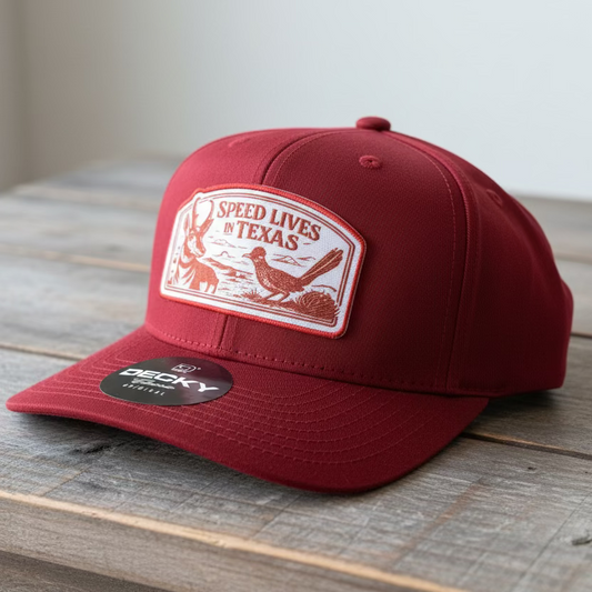 A red snapback cap with a full color embroidery patch that says "Speed Lives in Texas" with a pronghorn and roadrunner on a grey table background