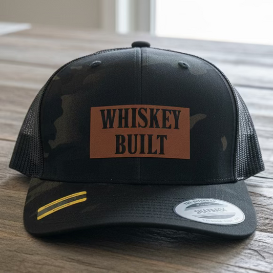 A black camo trucker snapback with a faux leather patch that says "Whiskey Built" on a rustic style table with grey neutral colors.