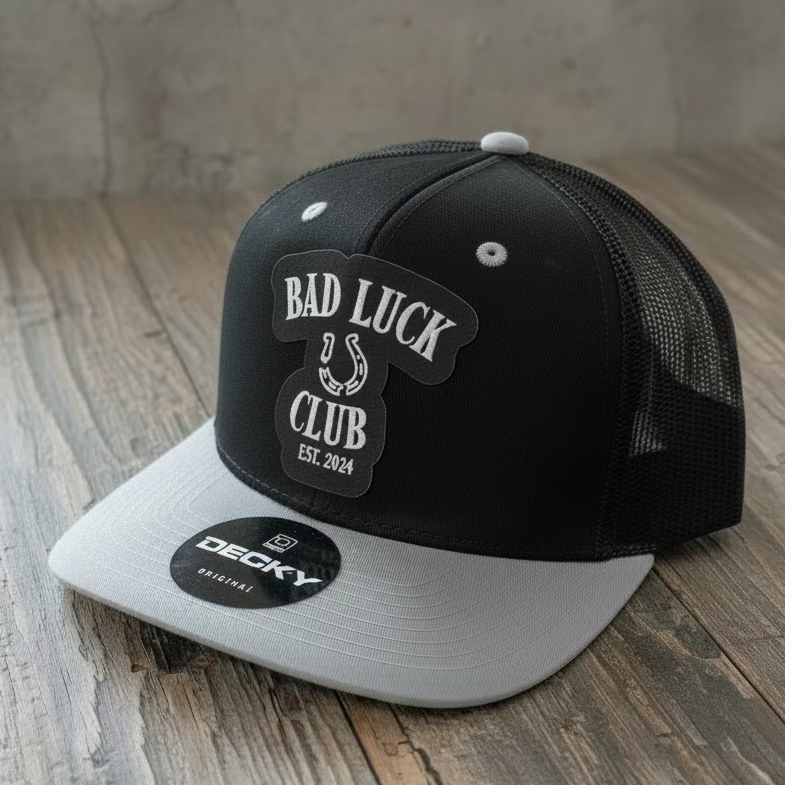 A black trucker snapback with a silver/grey bill and a black faux leather patch that says "Bad Luck Club Est. 2024" in silver with a silver broken horseshoe, on a rustic style table in grey neutral colors