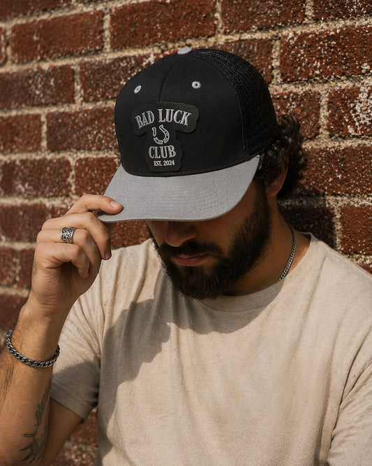 Man wearing a black trucker snapback with a silver/grey bill, with a black faux leather patch that says "Bad Luck Club Est. 2024"  in silver against a brick wall.