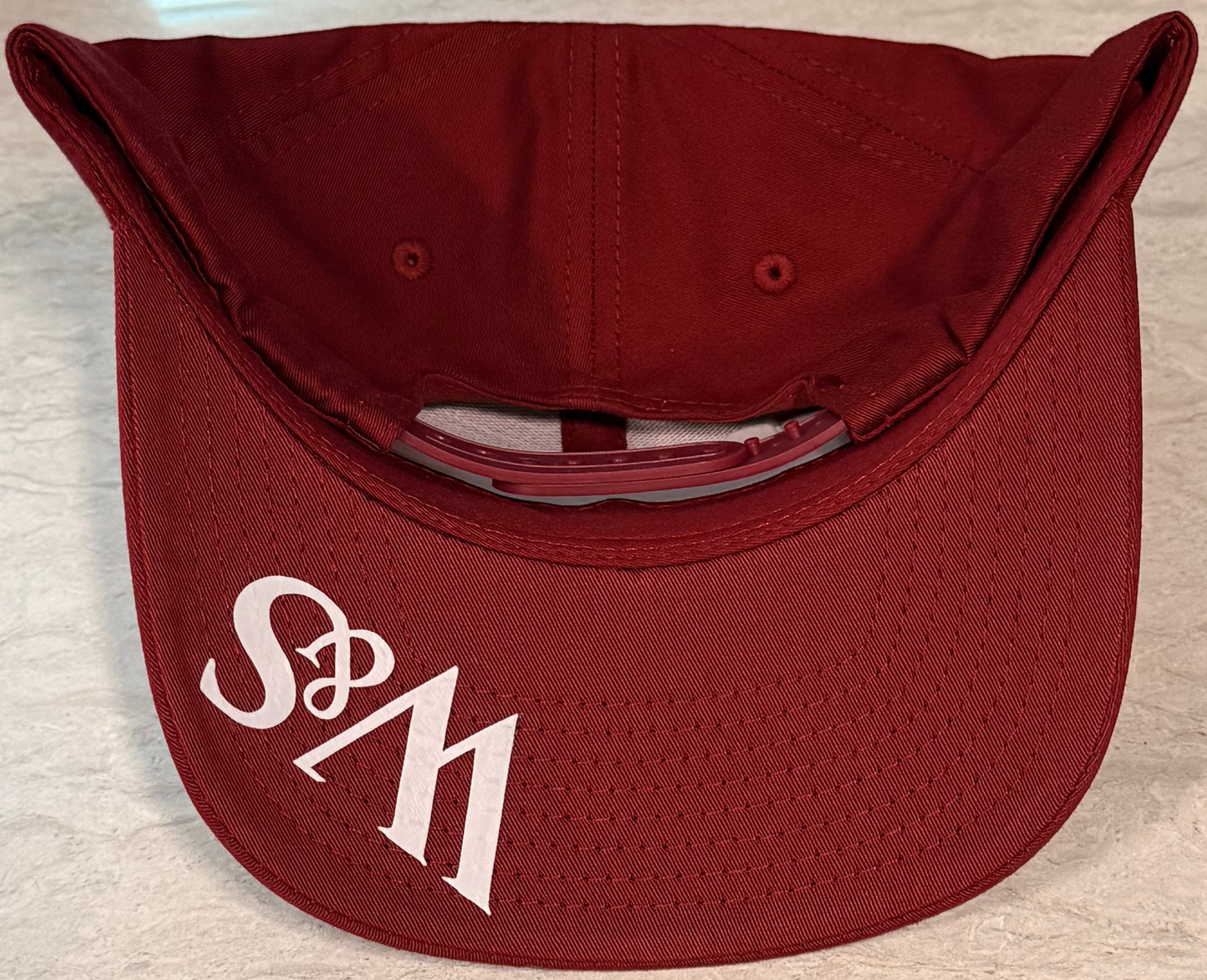 The underneath of a wine colored snapback with the logo W&S in white on the bill against a grey neutral background
