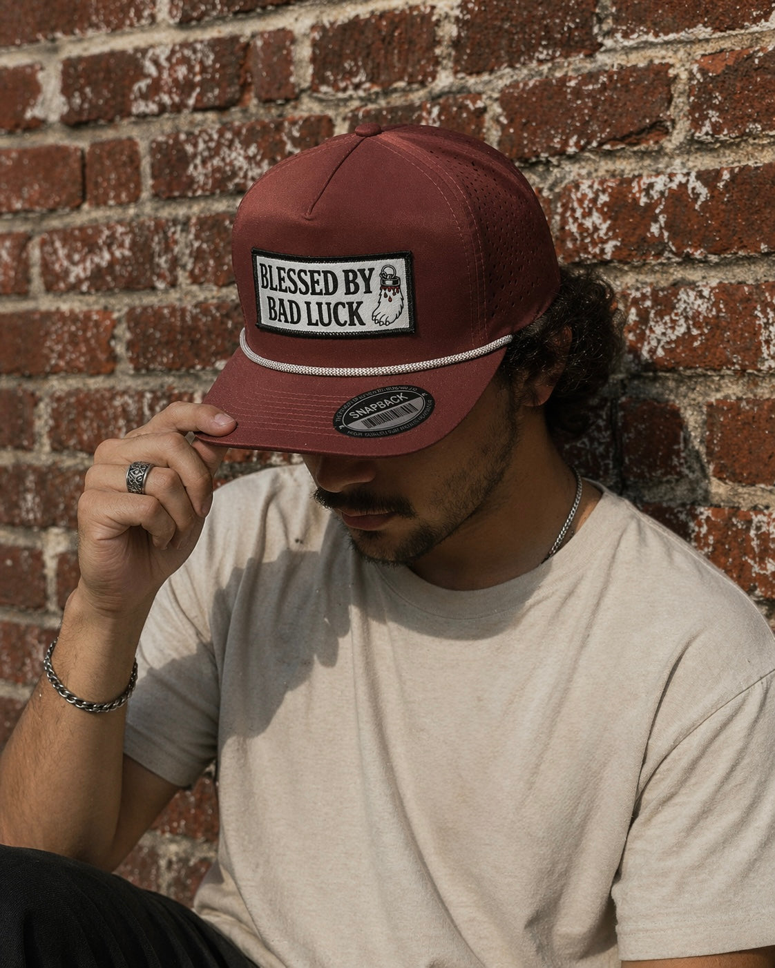 A man wearing a Wine colored roped sport laser mesh snapback with a full color embroidery patch that says "Blessed By Bad Luck" with a bloody lucky rabbits foot against a red brick background