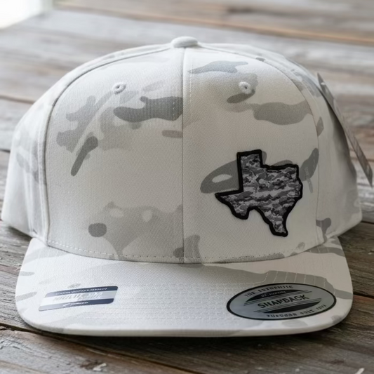 A white camo snapback with a white camo 3D embroidery patch of state Texas Shape/Pride off centered on a rustic style table with neutral colors