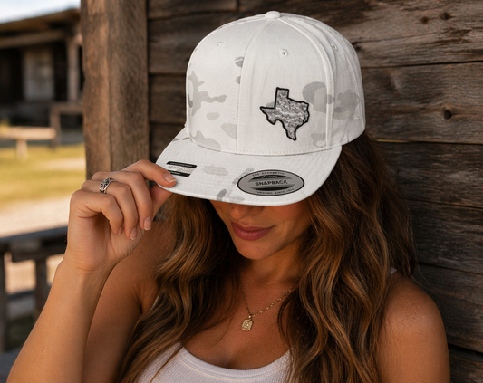 a women wearing a 6 panel snapback white camo hat that has a 3D embroidery patch of the state of Texas with white camo design against rustic wooden walls.