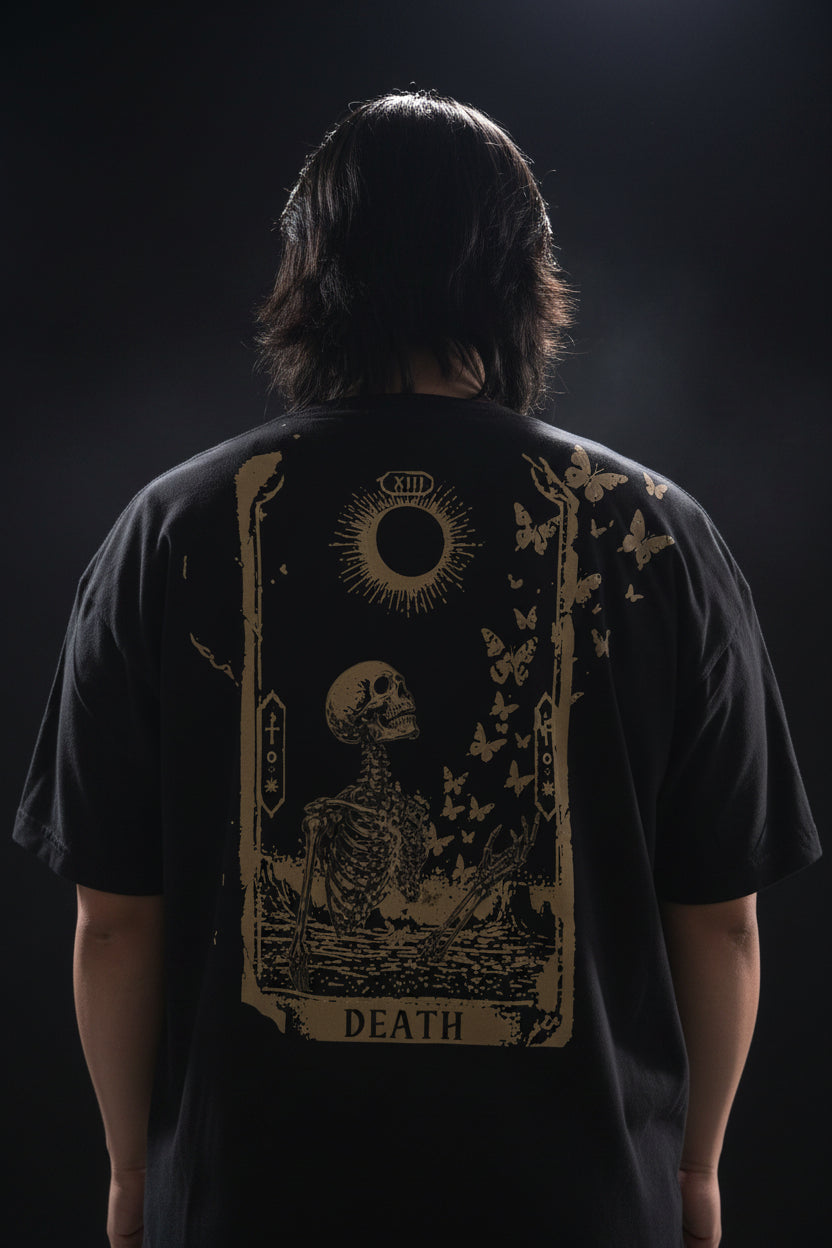 A person wearing a black t-shirt with a gold design of a skeleton turning into butterflies with a radiant new moon and tarot border