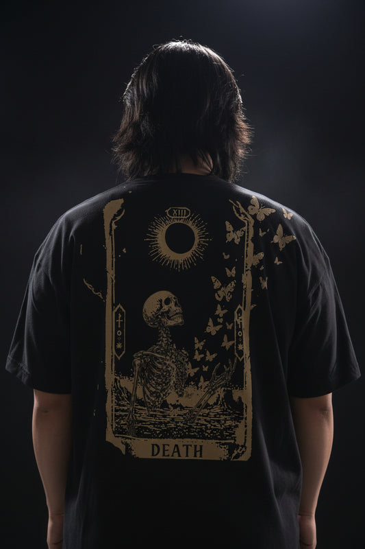 A person wearing a black t-shirt with a gold design of a skeleton turning into butterflies with a radiant new moon and tarot border