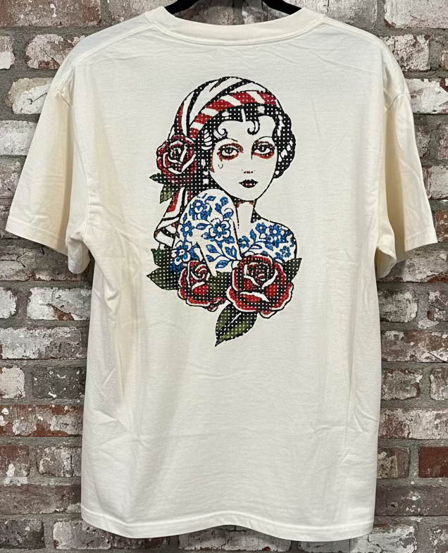 An ivory shirt that has an image of a traditional tattoo inspired Gypsy Girl, against a grey brick background