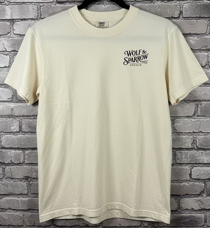 An ivory t-shirt with the logo Wolf & Sparrow Studio in black against a grey brick background