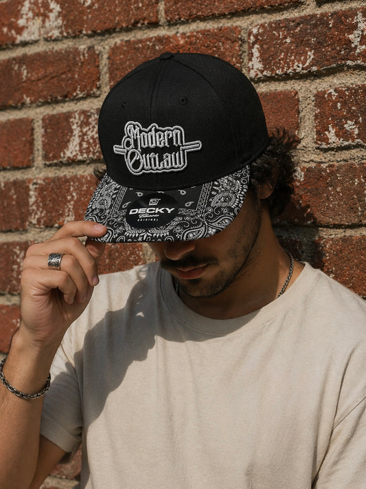 A man wearing a black snapback with a black and white paisley bill and a 3D embroidery patch that says "Modern Outlaw" against a red brick background.