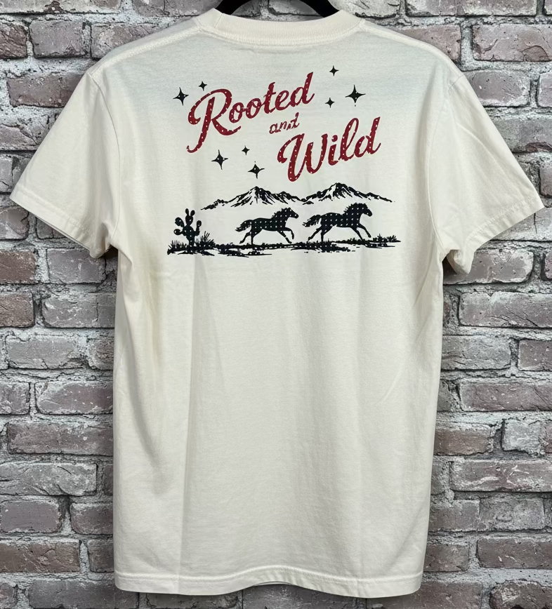  an ivory shirt that says Rooted and Wild with a mountain range, horses, and cactus against a grey brick background