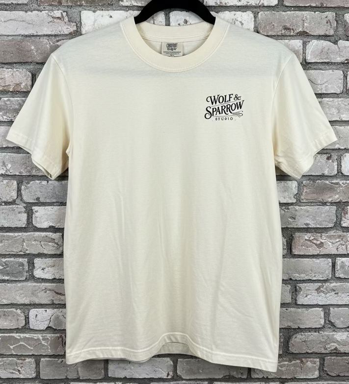 The front of an ivory shirt that has the logo Wolf & Sparrow Studio in black on the breast against a grey brick background