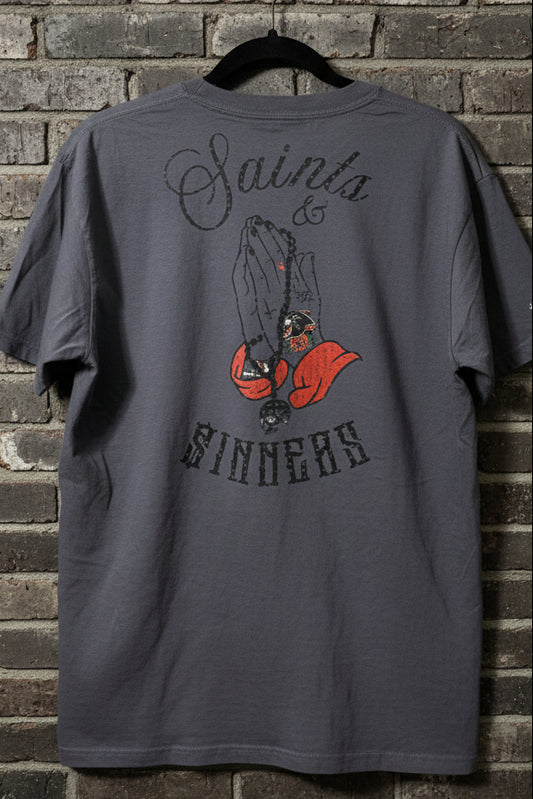 The back of a graphite t-shirt with the words "Saints & Sinners" with tattooed praying hands and a skull rosary against a grey brick background