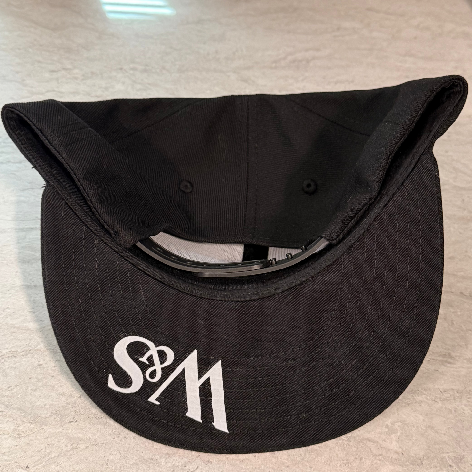 The underside of a black snapback with the logo W&S in white on the black bill
