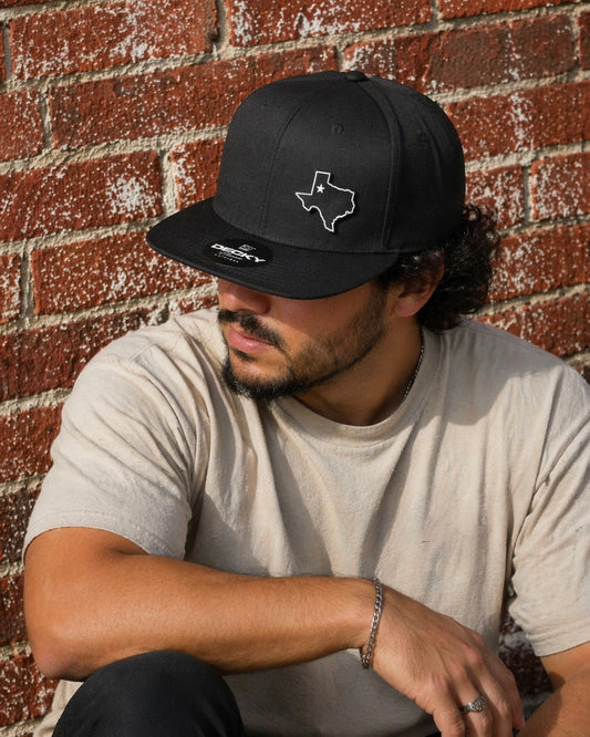A man wearing a black snapback with a PVC patch in the shape of the state Texas with a white outline against red brick