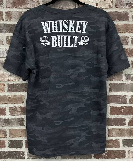 A black camo shirt that says "Whiskey Built" with spilling shot glasses in white against a grey brick background