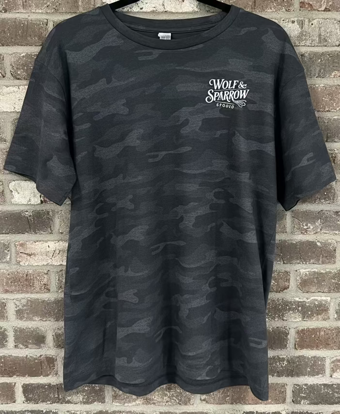 A black camo shirt with the logo Wolf & Sparrow Studio in white against a grey rustic wall