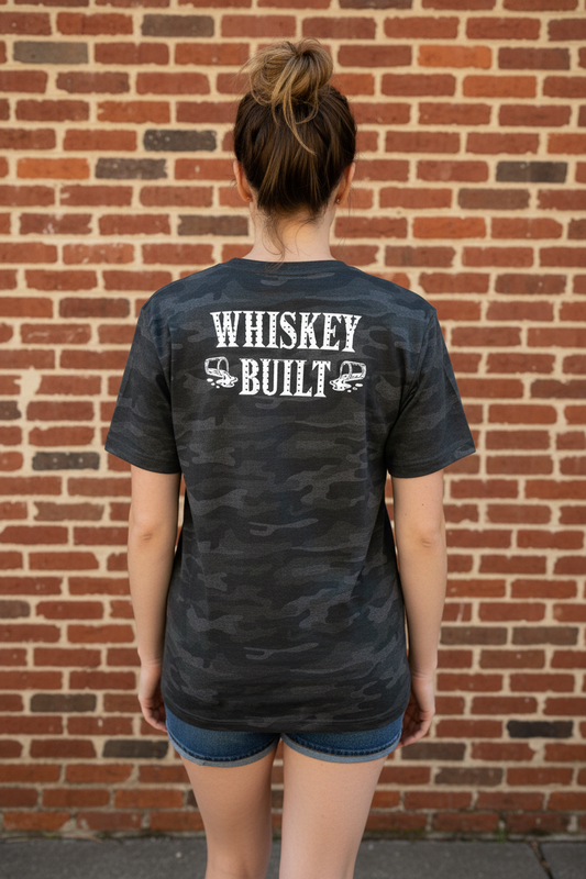 A woman wearing a black camo shirt that says "Whiskey Built" with spilling shot glasses in white against a brick background