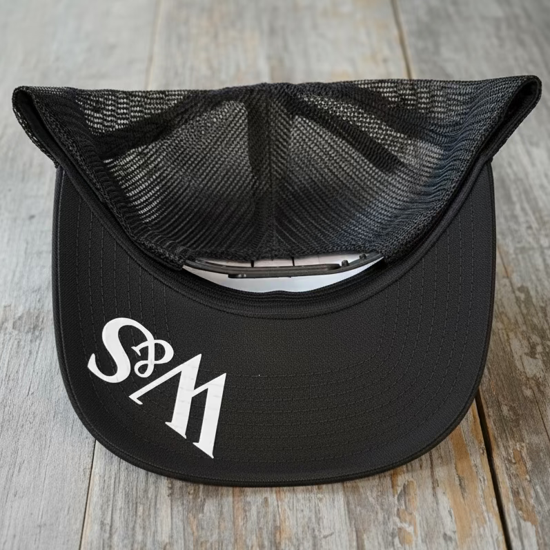 The underside of a black camo mesh trucker snapback with the logo W&S in white on the bill