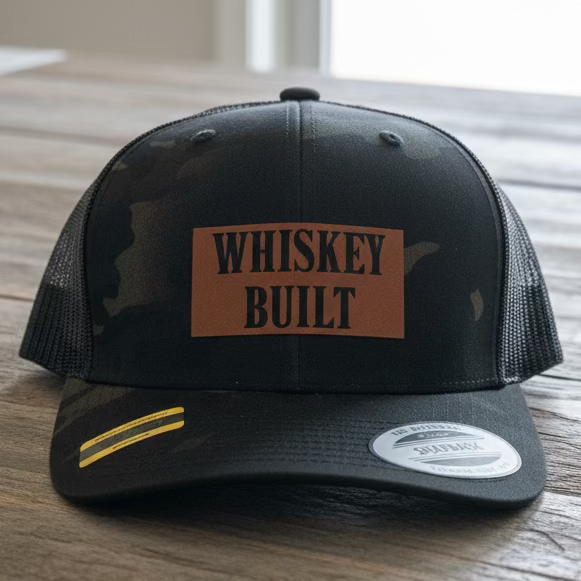 A black camo trucker snapback with a faux leather patch that says "Whiskey Built" on a rustic style table with grey neutral colors.