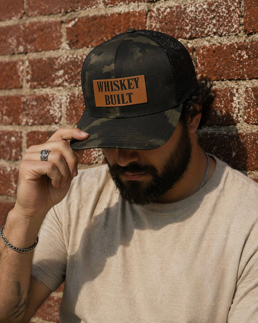 Man wearing a snapback with a faux leather patch that says 'Whiskey Built', against a brick wall